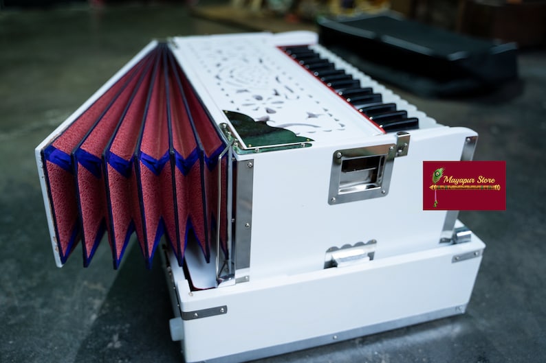 May include: A white harmonium with a red and blue bellows. The harmonium has a carved wooden design and a silver trim. The harmonium is open and the bellows are extended. The harmonium is sitting on a grey floor. Mayapur Store is printed on a red label.