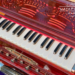 Pode incluir: Um harm&oacute;nio de madeira vermelho escuro com um teclado branco e entalhes florais intrincados no topo. O harm&oacute;nio tem um forro de feltro vermelho e uma placa de nome de lat&atilde;o que diz "MAYAPUR STORE HARMONIUM".