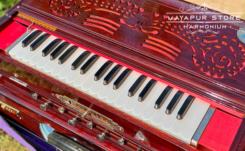 Pode incluir: Um harm&oacute;nio de madeira vermelho escuro com um teclado branco e entalhes florais intrincados no topo. O harm&oacute;nio tem um forro de feltro vermelho e uma placa de nome de lat&atilde;o que diz "MAYAPUR STORE HARMONIUM".