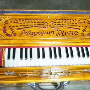 K&ouml;nnte beinhalten: Ein Harmonium aus Holz mit einem roten und goldenen Design. Das Harmonium hat eine Tastatur mit wei&szlig;en und schwarzen Tasten und den Text "Manyapur Stone" ist in die Oberseite geschnitzt.