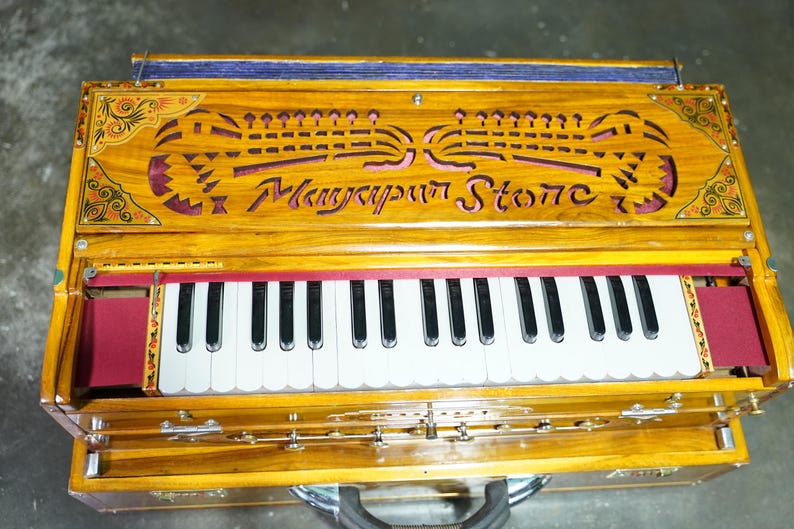 K&ouml;nnte beinhalten: Ein Harmonium aus Holz mit einem roten und goldenen Design. Das Harmonium hat eine Tastatur mit wei&szlig;en und schwarzen Tasten und den Text "Manyapur Stone" ist in die Oberseite geschnitzt.
