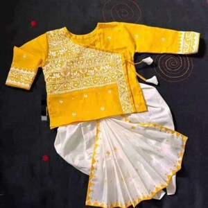 May include: A yellow and white traditional outfit. The top is yellow with white floral embroidery and three-quarter sleeves. The bottom is white with a yellow patterned border. The outfit is likely for a child.