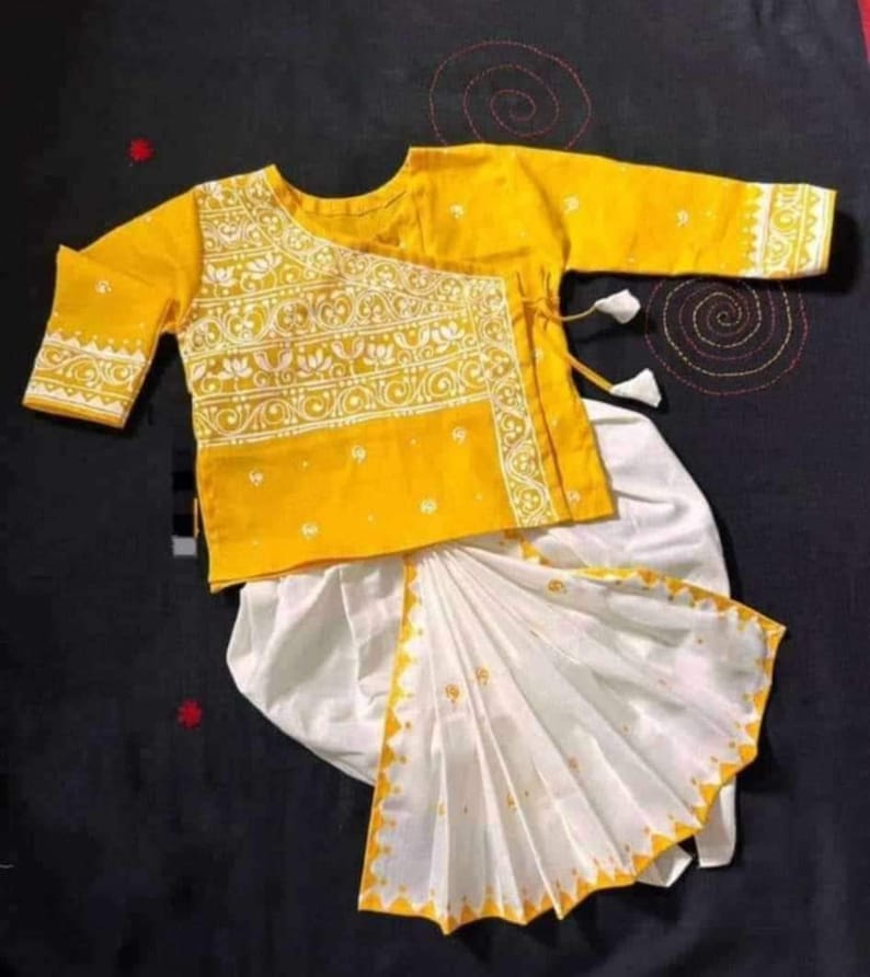 May include: A yellow and white traditional outfit. The top is yellow with white floral embroidery and three-quarter sleeves. The bottom is white with a yellow patterned border. The outfit is likely for a child.