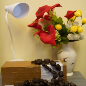 K&ouml;nnte beinhalten: Eine braune Holzperlenkette mit einer wei&szlig;en Lampe und einer Vase mit roten und gelben Blumen im Hintergrund.