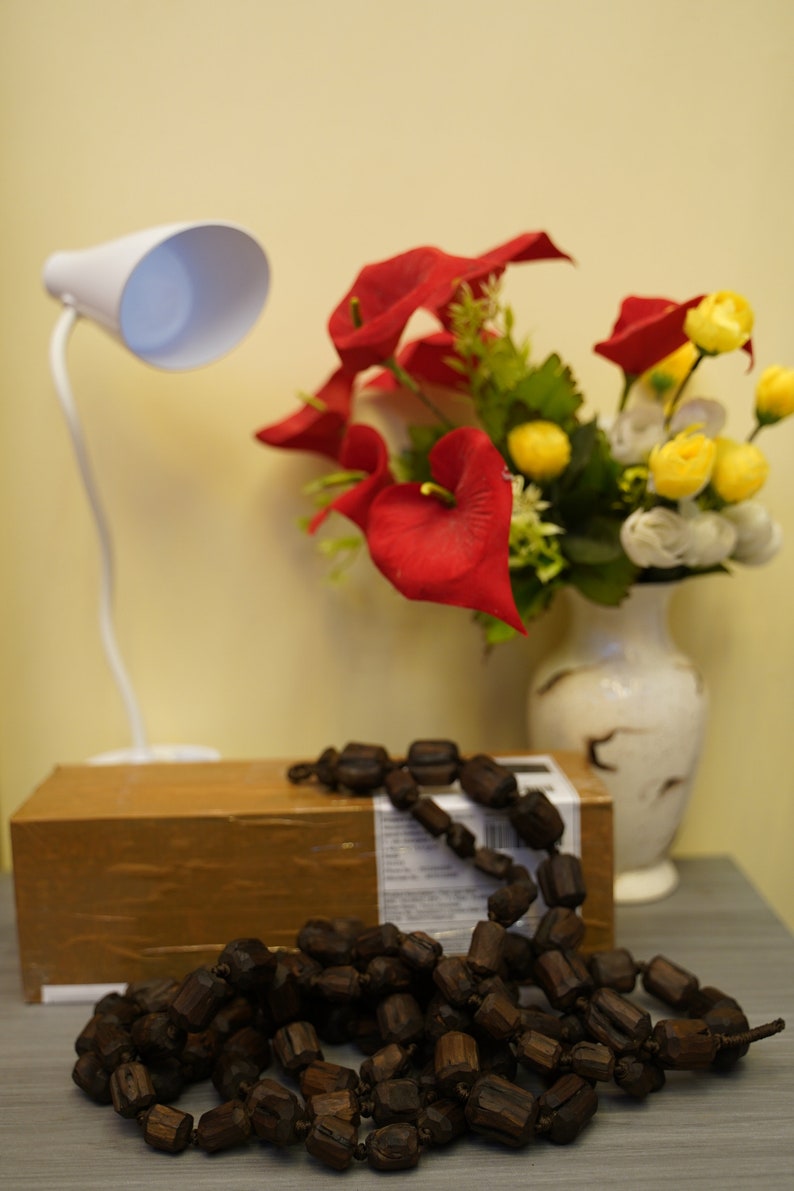 K&ouml;nnte beinhalten: Eine braune Holzperlenkette mit einer wei&szlig;en Lampe und einer Vase mit roten und gelben Blumen im Hintergrund.
