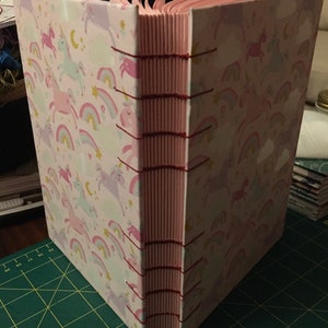 May include: A pink and white book with a unicorn and rainbow pattern. The book is bound with red thread and has a pink spine.