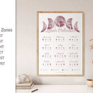 May include: A printable lunar calendar for 2023 featuring a pink watercolour moon design. The calendar shows the moon phases for each month, with the dates of the new moon, first quarter, full moon, and last quarter. The calendar is framed and displayed on a table with a vase of dried flowers.