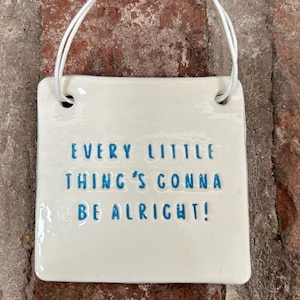 Könnte beinhalten: Ein weißes quadratisches Schild aus Keramik mit einer blauen Aufschrift, die "Every little thing's gonna be alright!" lautet, hängt an einer weißen Schnur.