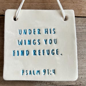 Könnte beinhalten: Quadratische, weiße Keramikplatte mit dem Text "UNDER HIS WINGS YOU FIND REFUGE. PSALM 91:4" in blauer Schrift. Die Platte hat zwei Löcher oben und eine weiße Schnur zum Aufhängen.