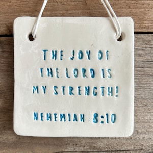 May include: White ceramic square plaque with blue text that reads "The joy of the Lord is my strength! Nehemiah 8:10". The plaque has two holes at the top for hanging.