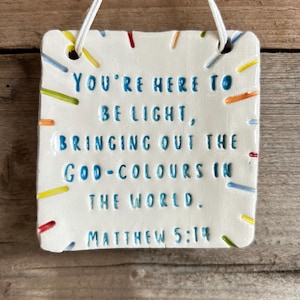 May include: A white ceramic plaque with the text "YOU'RE HERE TO BE LIGHT, BRINGING OUT THE GOD-COLOURS IN THE WORLD. MATTHEW 5:14" in blue lettering. The plaque is decorated with colourful dashes and has a white string for hanging.