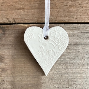 Puede incluir: Adorno en forma de corazón de cerámica blanca con un diseño floral en relieve. Se adjunta una cinta blanca transparente para colgar. El adorno se muestra sobre un fondo de madera rústica.
