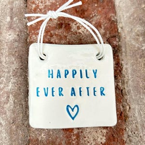 May include: White ceramic sign with a blue inscription that reads "Happily Ever After" and a blue heart. The sign has two holes at the top and is attached to a white string.