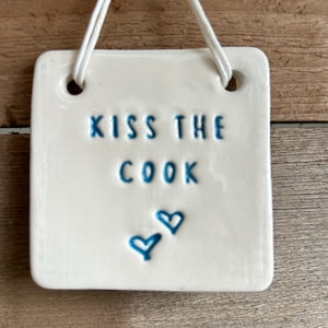 Op de afbeelding: Een wit keramisch bord met blauwe tekst die "Kiss the Cook" luidt en twee blauwe hartjes. Het bord heeft twee gaten bovenaan om het op te hangen.