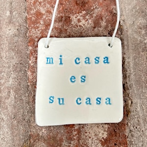 Könnte beinhalten: Ein weißes Keramik-Schild mit blauem Text, auf dem "mi casa es su casa" steht.