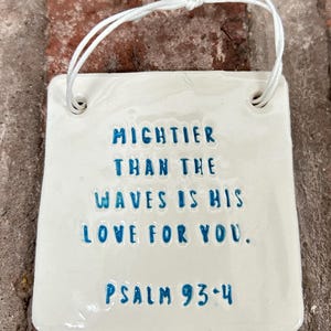 Könnte beinhalten: Weiße Keramikfliese mit blauer Beschriftung, die "MICHTIER THAN THE WAVES IS HIS LOVE FOR YOU. PSALM 93:4" lautet. Die Fliese hat oben ein Loch zum Aufhängen.