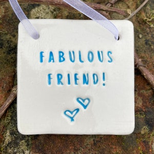 Può includere: Piastrella di ceramica bianca con la scritta "Fabulous Friend!" in blu e due cuori blu.