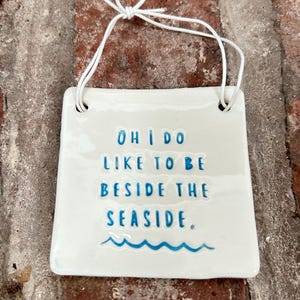 Może przedstawiać: Biała ceramiczna tabliczka z niebieskim tekstem, który brzmi "Oh I do like to be beside the seaside." Tabliczka ma sznurek do zawieszenia.