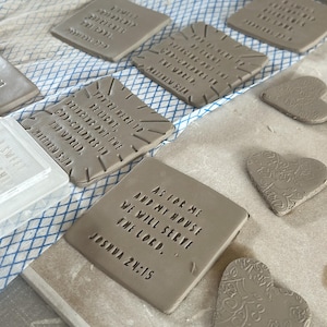 May include: Handmade clay tiles and heart-shaped ornaments are displayed. The rectangular tiles have embossed text, including the phrase "As for me and my house, we will serve the Lord." The hearts have a decorative floral pattern.
