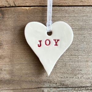 May include: White ceramic heart ornament with a red "JOY" inscription. The ornament has a hole at the top for hanging.