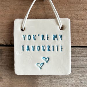 Op de afbeelding: Witte keramische tegel met een blauwe inscriptie die "You're my favourite" luidt, met twee blauwe harten onder de tekst. De tegel hangt aan een wit touwtje.