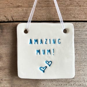 May include: White ceramic plaque with the text "AMAZING MUM!" in blue lettering and two blue hearts.