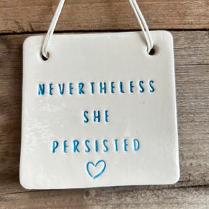 Può includere: Un cartello quadrato in ceramica bianca con scritta blu che recita "Nevertheless she persisted" con un cuore blu in basso.