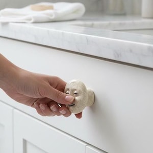 May include: A cream-colored cabinet knob with a smiling face design. The knob is attached to a white cabinet door. The knob has a textured surface and is made of ceramic. The bathroom cabinet has a marble countertop.