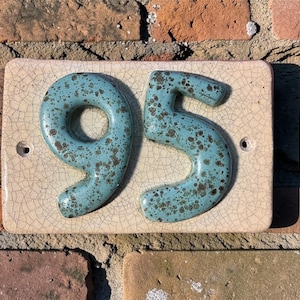 May include: A rectangular ceramic house number plaque featuring the digits 95 in a speckled teal glaze. The background tile is a light beige with a visible crackle finish. Two circular mounting holes are positioned on the left and right sides.