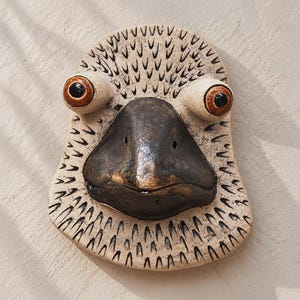 Handmade ceramic bird wall mask with speckled cream surface, carved black feather marks, oversized brown eyes, and a dark sculpted beak on a warm plaster wall.