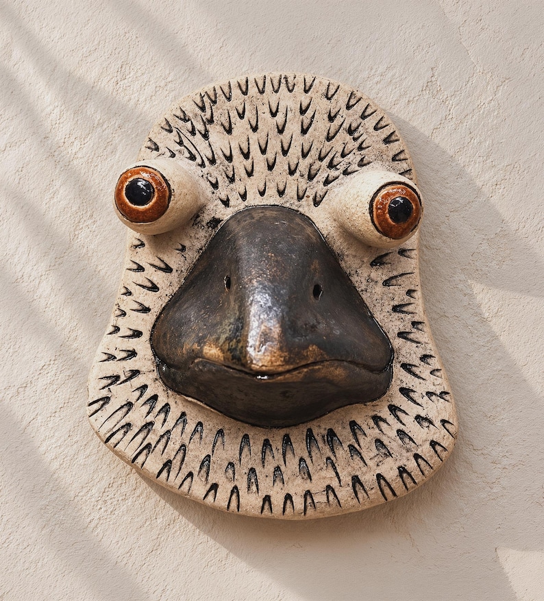 Handmade ceramic bird wall mask with speckled cream surface, carved black feather marks, oversized brown eyes, and a dark sculpted beak on a warm plaster wall.