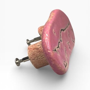 Könnte beinhalten: Ein rosa Keramik-Schrankknopf mit strukturierter Oberfläche und dunklen Craquelé-Details. Der Knopf ist an einer hellbraunen Holzbasis mit einer silbernen Schraube befestigt. Diese dekorative Hardware verleiht Möbeln eine einzigartige Note.