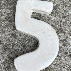 Op de afbeelding: Een keramisch cijfer vijf in een cr&egrave;mewitte kleur. Het cijfer heeft een licht verweerde uitstraling met enkele onvolkomenheden. Het cijfer is geplaatst op een gestructureerd, grijs betonnen oppervlak.