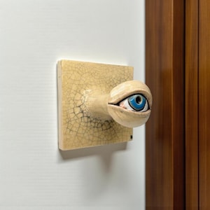 Könnte beinhalten: Keramik-Türgriff mit Augendesign. Das Auge hat eine blaue Iris und ist auf einer beige, rissigen quadratischen Basis angebracht. Der Griff ist an einer weißen Wand montiert, mit einem sichtbaren Holzrahmen.
