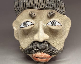 May include: A ceramic face sculpture with a mustache, eyebrows, and a textured surface. The sculpture features a light beige face with black and gray details, including a striped hair design and a red-orange mouth.