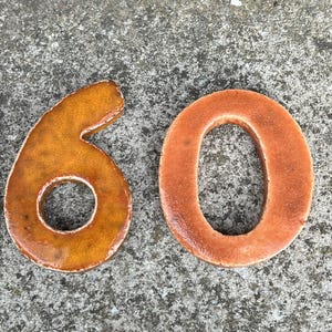 Op de afbeelding: Keramisch nummer 60 in een warm, aards kleurenpalet. Het cijfer 6 is een rijk, goudbruin, terwijl de 0 een gedempt, roodbruin is. De cijfers staan op een getextureerd, grijs oppervlak.