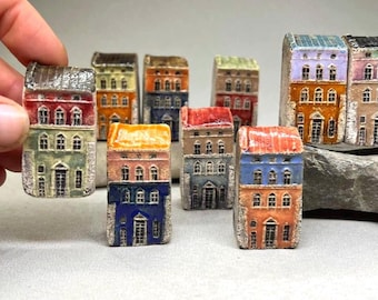 May include: A collection of small ceramic houses, each painted in different colours such as red, orange, and blue. The houses have multiple storeys with detailed windows and doors. They are displayed on a light grey surface.