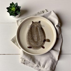 Puede incluir: Plato de cerámica redondo, blanquecino, con un diseño de animal pintado a mano en marrón y beige. El plato descansa sobre una servilleta de lino beige. Una pequeña planta suculenta en maceta está en el fondo.