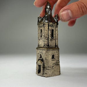 May include: A miniature ceramic church tower with a detailed, textured exterior. The tower is primarily beige with darker brown accents on the roof and trim. The tower has arched windows and a small door.