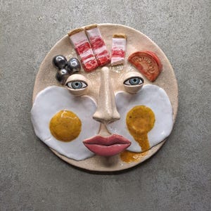 May include: A ceramic plate with a face design featuring fried eggs, bacon, and a tomato slice. The face includes blue-eyed eyeballs, a nose, and pink lips. The plate has a beige, cracked finish.