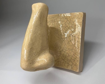 May include: A ceramic nose sculpture in a light beige color, attached to a square, textured tile. The nose has a detailed, realistic shape, and the tile has a cracked, aged appearance. The artwork is likely for home decor.