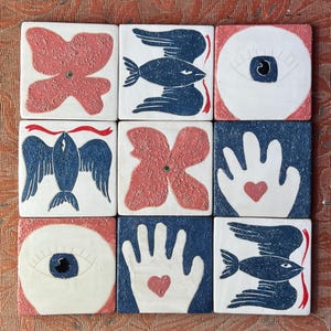May include: Nine square ceramic tiles with various designs. The tiles feature red and blue illustrations of a butterfly, fish, eye, hand with a heart, and a flower. The tiles are arranged in a 3x3 grid on a textured surface.