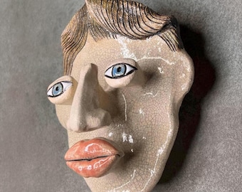 May include: A ceramic wall mask featuring a stylized human face with a cream-colored crackle glaze finish. The sculpture displays brown textured hair, two prominent blue eyes, a large protruding nose, and full, glossy peach-colored lips.