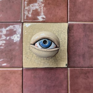 May include: A ceramic eye sculpture with a blue iris and tan eyelid is centered on a square, light yellow tile. The tile is surrounded by reddish-brown tiles. The eye is a unique decorative piece.