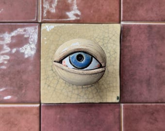 May include: A ceramic eye sculpture with a blue iris and tan eyelid is centered on a square, light yellow tile. The tile is surrounded by reddish-brown tiles. The eye is a unique decorative piece.