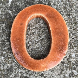Op de afbeelding: Een witte keramische mok met een zwart handvat en de tekst "But First, Coffee" in een vetgedrukte, zwarte letter. De mok is gevuld met een donkere vloeistof, waarschijnlijk koffie, en staat op een lichtgekleurd oppervlak.