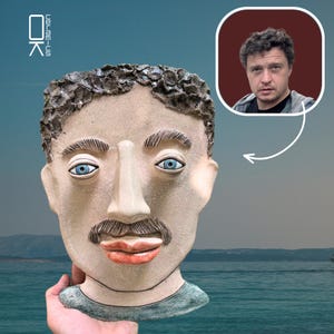 May include: A ceramic sculpture of a man's face with blue eyes, a mustache, and a textured hair design. The sculpture is held in a hand. A photo of a man is in the upper right corner. The background is a seascape.