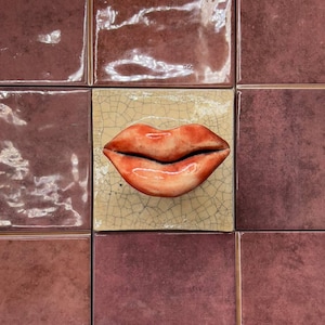 May include: A square tile with a pair of red lips in the center, surrounded by other square tiles in shades of burgundy. The lips are a glossy red with a black outline, set against a beige, cracked background.