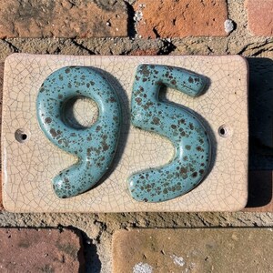 Op de afbeelding: Een keramische huisnummerplaat met de nummers "95". De nummers zijn turquoise met bruine spikkels, gemonteerd op een rechthoekige, gebroken witte tegel met een craquel&eacute; glazuur. Op een rode bakstenen muur.