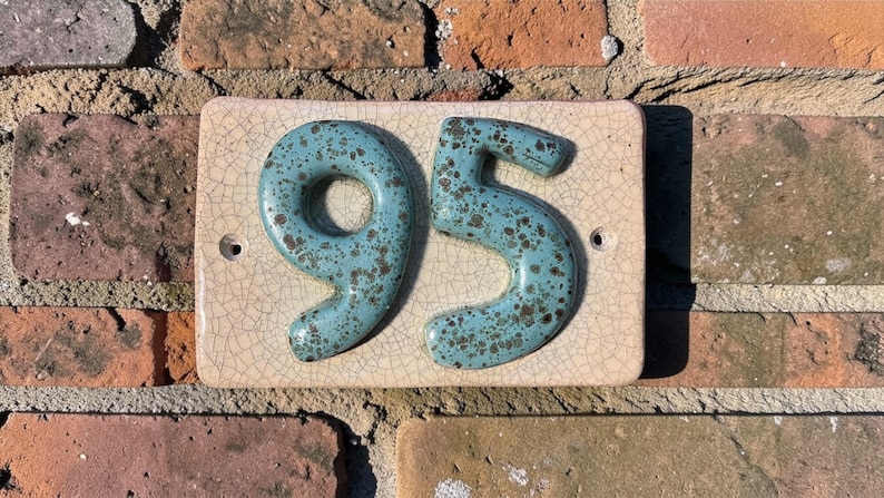 Op de afbeelding: Een keramische huisnummerplaat met de nummers "95". De nummers zijn turquoise met bruine spikkels, gemonteerd op een rechthoekige, gebroken witte tegel met een craquel&eacute; glazuur. Op een rode bakstenen muur.